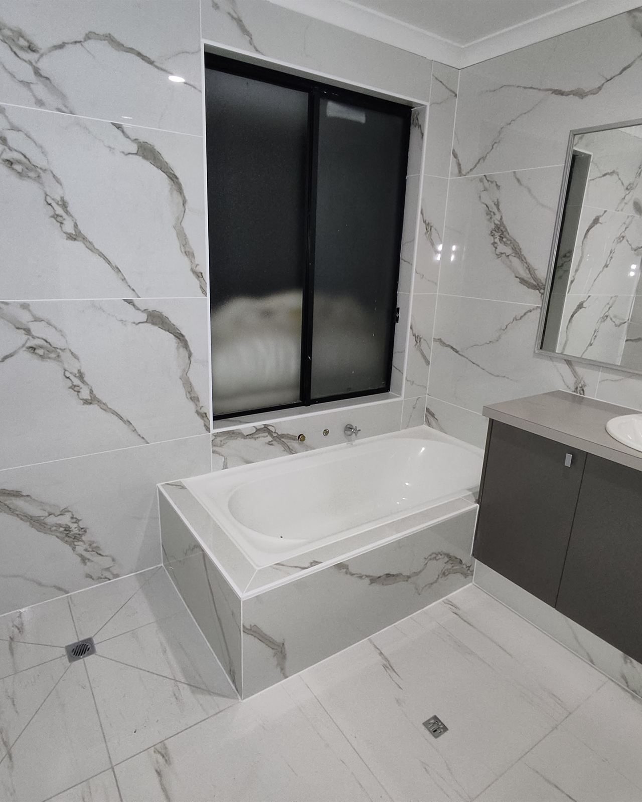 Bathroom tiled floor to ceiling in white marble-look tiles, with a tiled bath hob and dark vanity